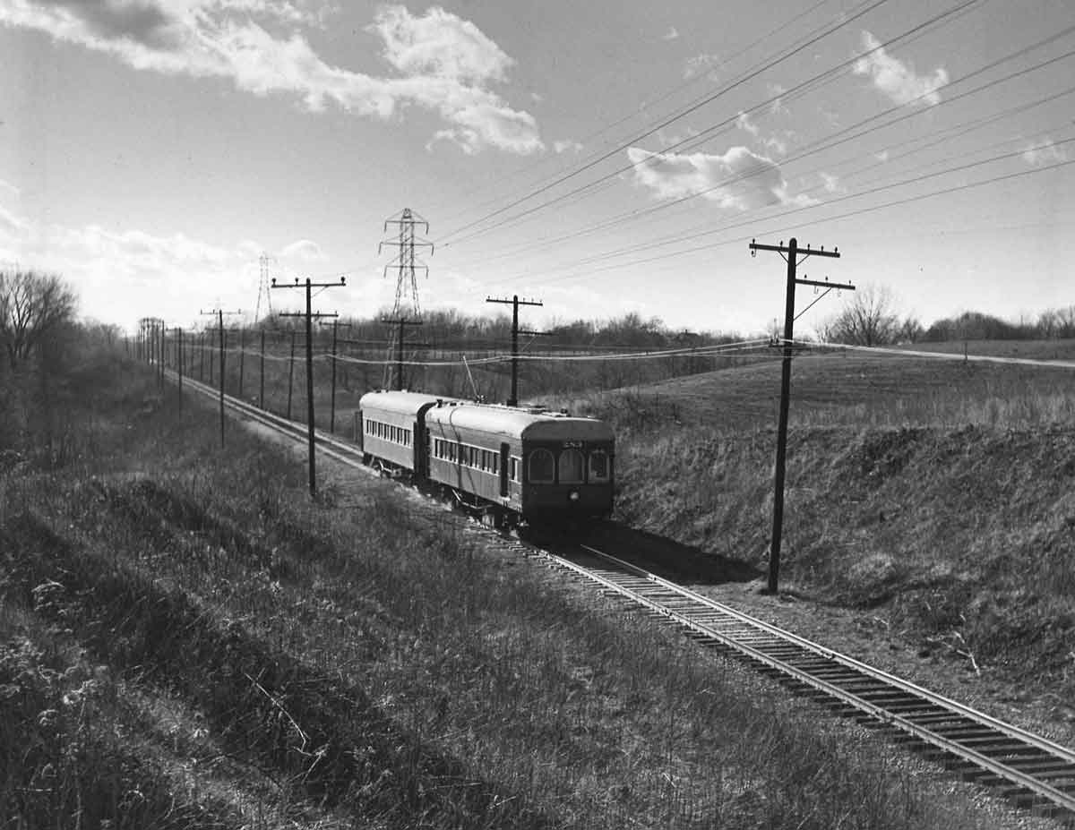 A black and white photo of a passenger train rolling down tracks near a hill