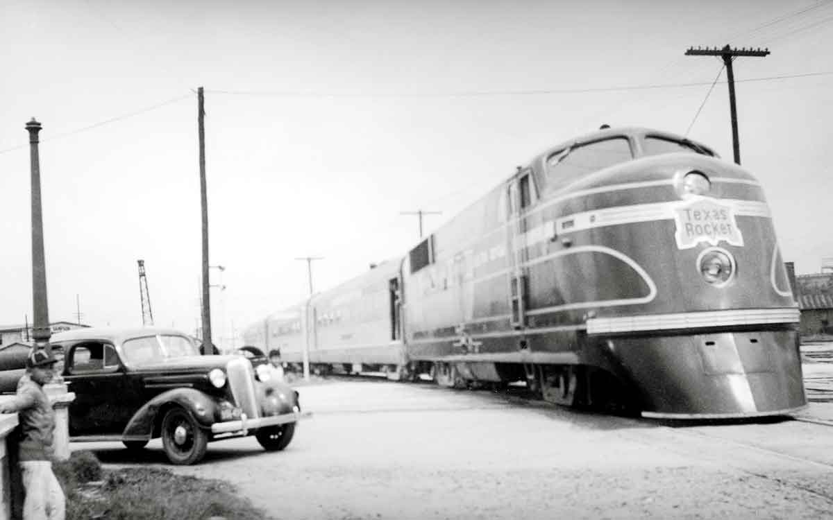 A black and white photo of a train passing by a car and young boy