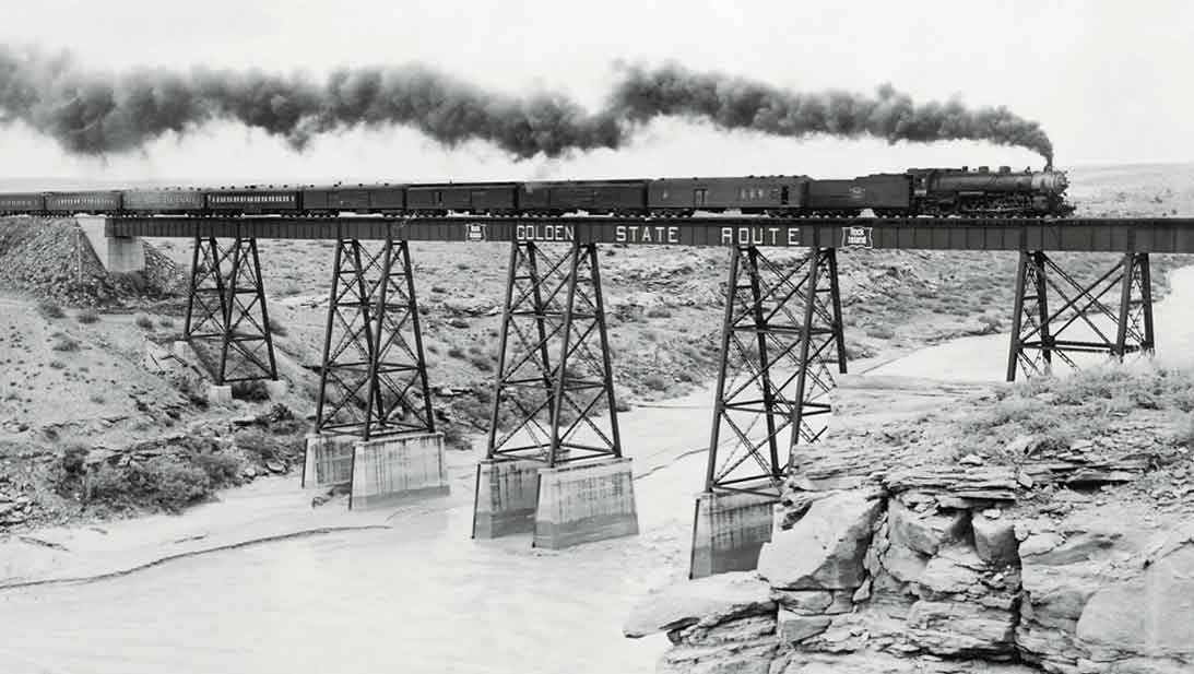 A black and white photo of a train with black smoke coming out of its chimney passing over a river