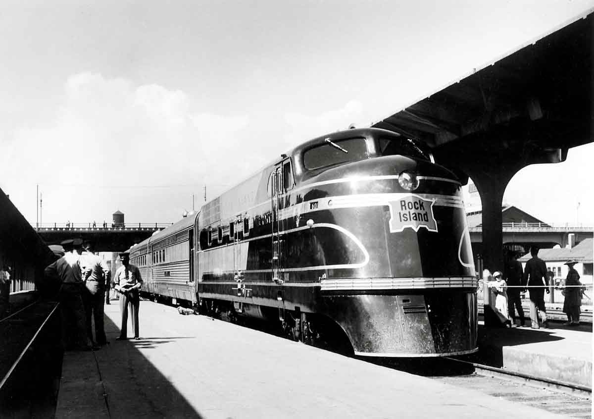 A black and white photo of a train parked at a station in front of passengers