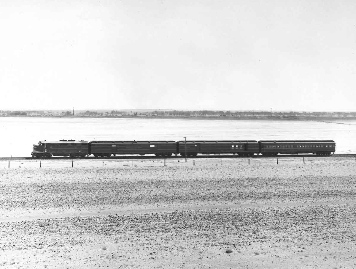 A black and white photo of a passenger train rolling through a flatland area