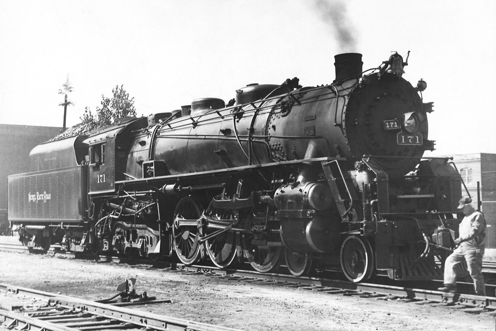A black and white photo of locomotive 4-6-4 stopped at the rail yard as a person walks across the tracks