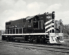 A black and white photo of a GP18 locomotive in a rail yard with the conductor looking or the window with his arm out the window