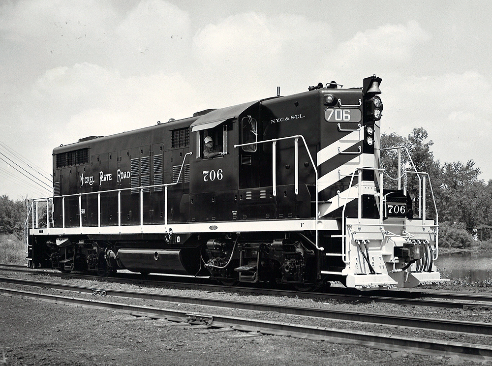 A black and white photo of a GP18 locomotive in a rail yard with the conductor looking or the window with his arm out the window