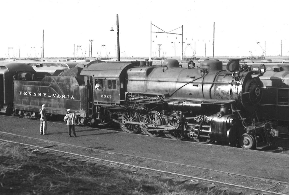 A black and white photo of a H9 2-8-0 Consolidation No. 3529 locomotive in a rail yard with two people talking next to it
