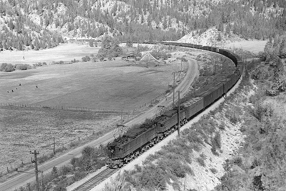 Two electric heavy freight motors pull a long freight train through a sparsely treed mountain scene.