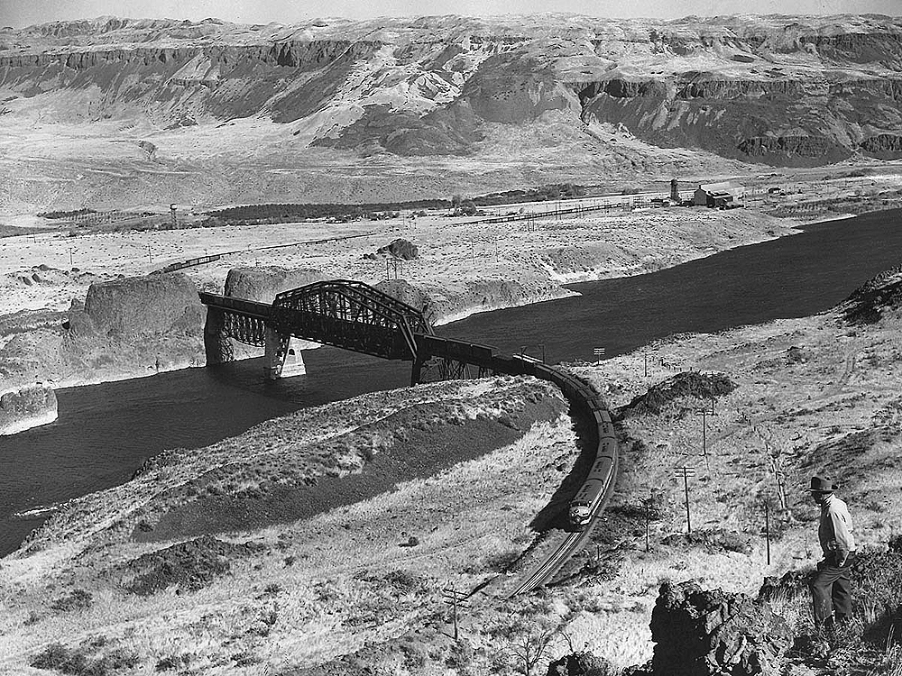 Wide scene of high-desert and mountains with a cab unit led train crossing a river.