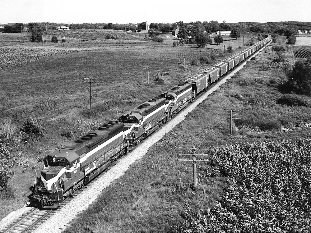 Mid-20th-century diesel locomotives lead a long train of covered hoppers in a rolling farm scene.