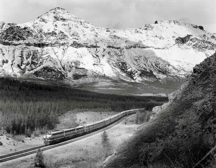 Cab-unit diesel locomotives lead a long passenger train on an s-curve at the foot of snow-capped mountains and forest.