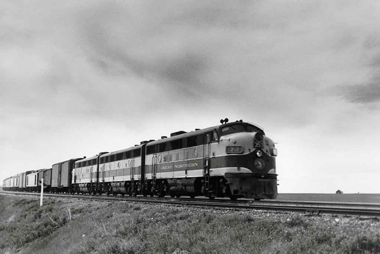 Cab unit diesel locomotives lead a mail-express train through a prairie.
