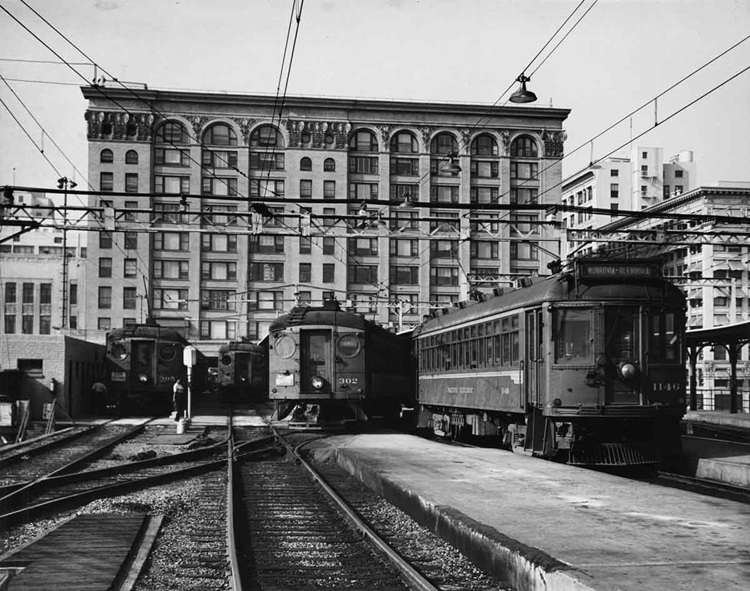 Passenger train terminal in an early 20th century city scene filled with streetcars or interurban cars.