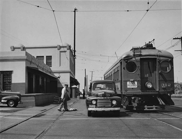 An interurban and an automobile appear stopped while a uniformed motorman walks to the cars with a stool in hand on a street.
