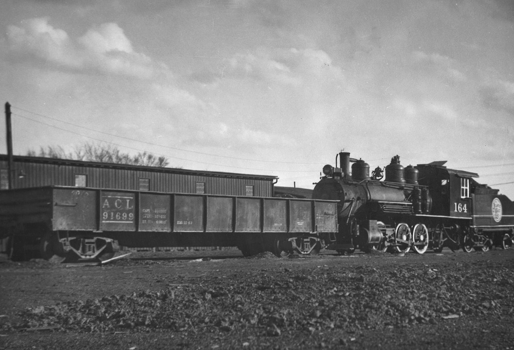 An 0-6-0 steam locomotive switcher with a gondola in a rail yard.