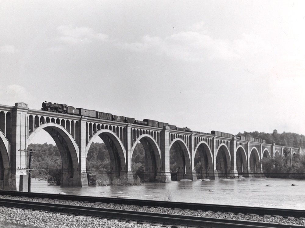 A steam locomotive leads a freight train over a river on a large stone or concrete arch bridge.
