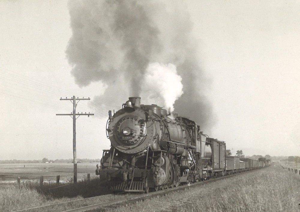 A steam locomotive leads a freight train up a long grade.