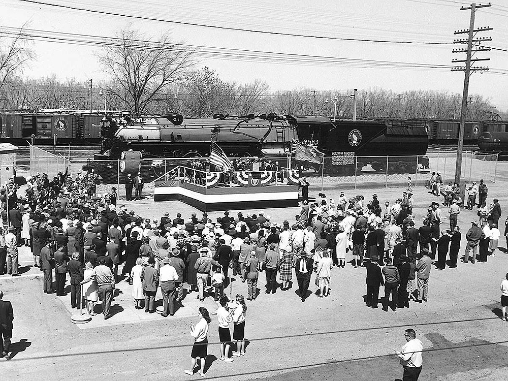 A crowd of people surrounds a platform and steam locomotive on display.