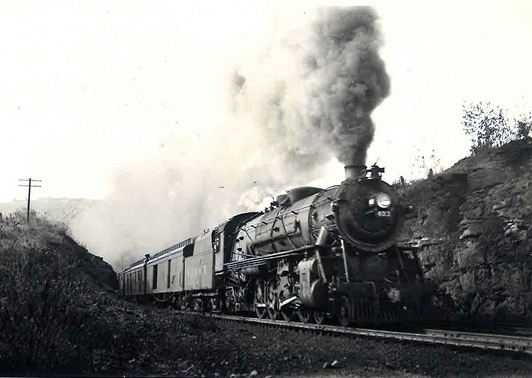 A steam locomotive leads a passenger train through a rock cut.