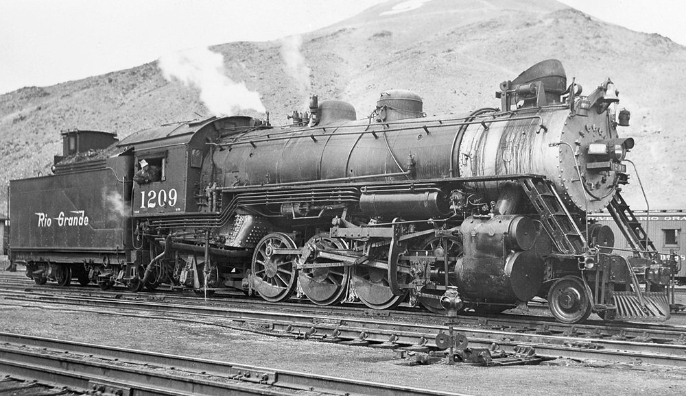 Side view of a 2-8-2 steam locomotive at the foot of an arid mountain.