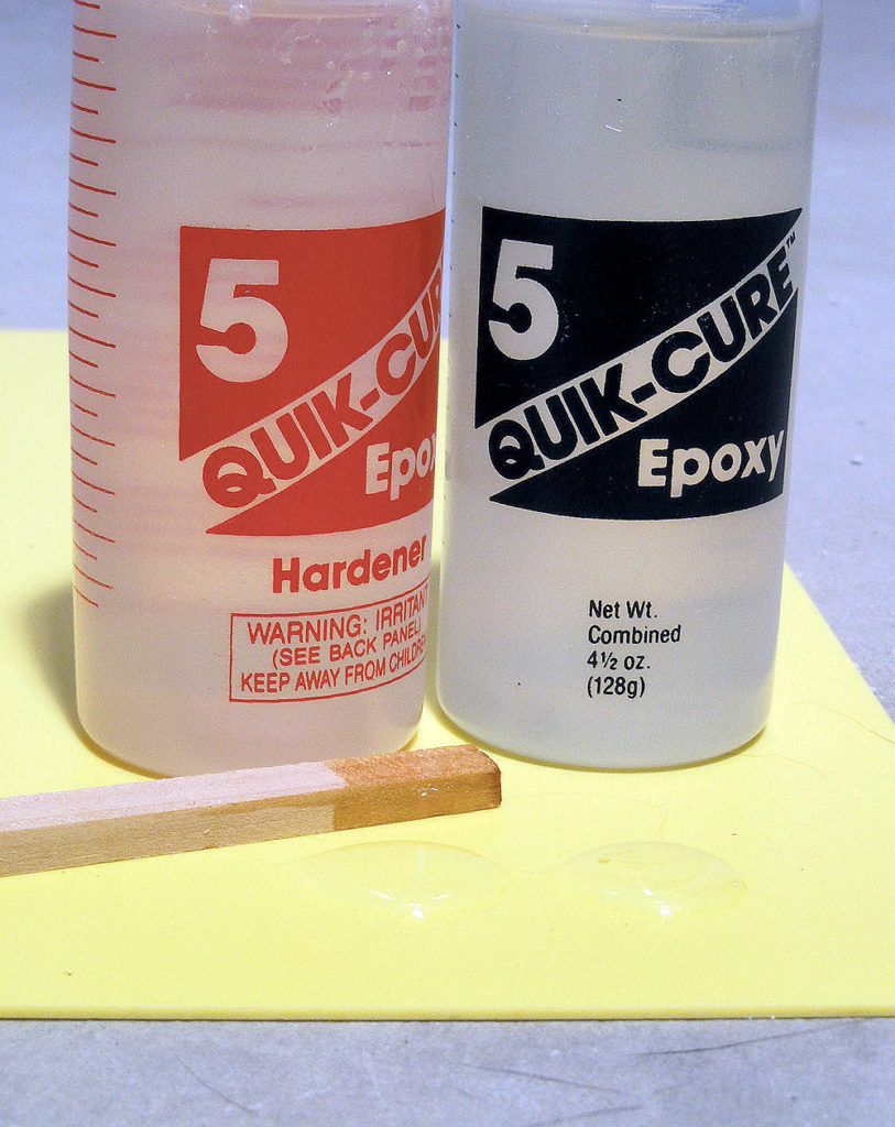 Bottles of two-part epoxy behind equal drops to be mixed with a stick.