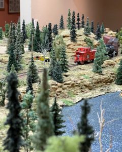 Photograph showing a river in the foreground, with a bright red caboose passing in the middle distance amid hills and sparse trees