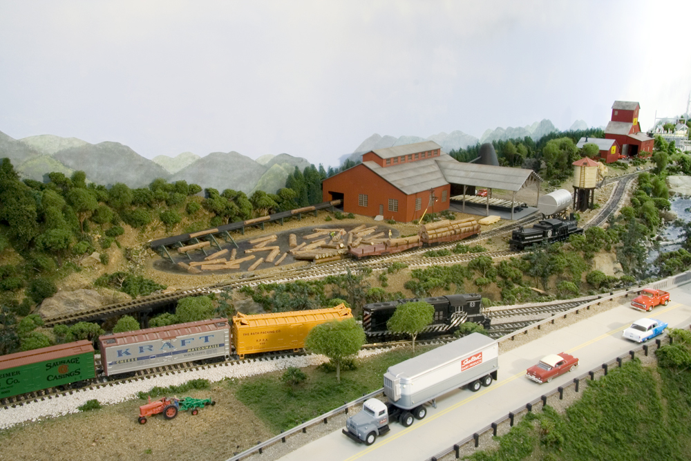 A diesel-locomotive-led Santa Fe train passes under a highway with a lumber mill in the background.