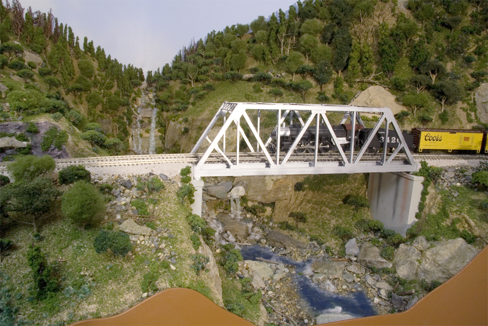 A double-tracked bridge hosts a steam-locomotive hauled Santa Fe train over a green-and-rock filled gorge.