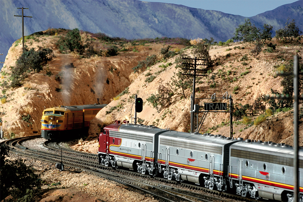 A Union Pacific train nears the top of the grade in an arid scene at "Summit" in Cajon Pass while a Santa Fe steam locomotive idles near the dispatcher's station.