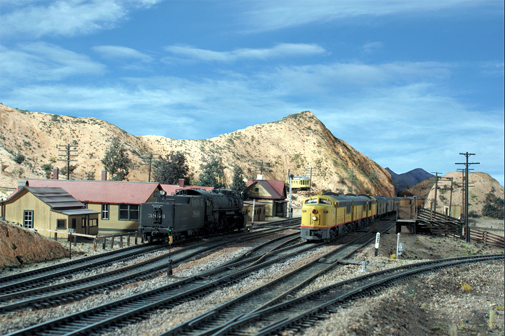 A Union Pacific train nears the top of the grade in an arid scene at "Summit" in Cajon Pass while a Santa Fe steam locomotive idles near the dispatcher's station.