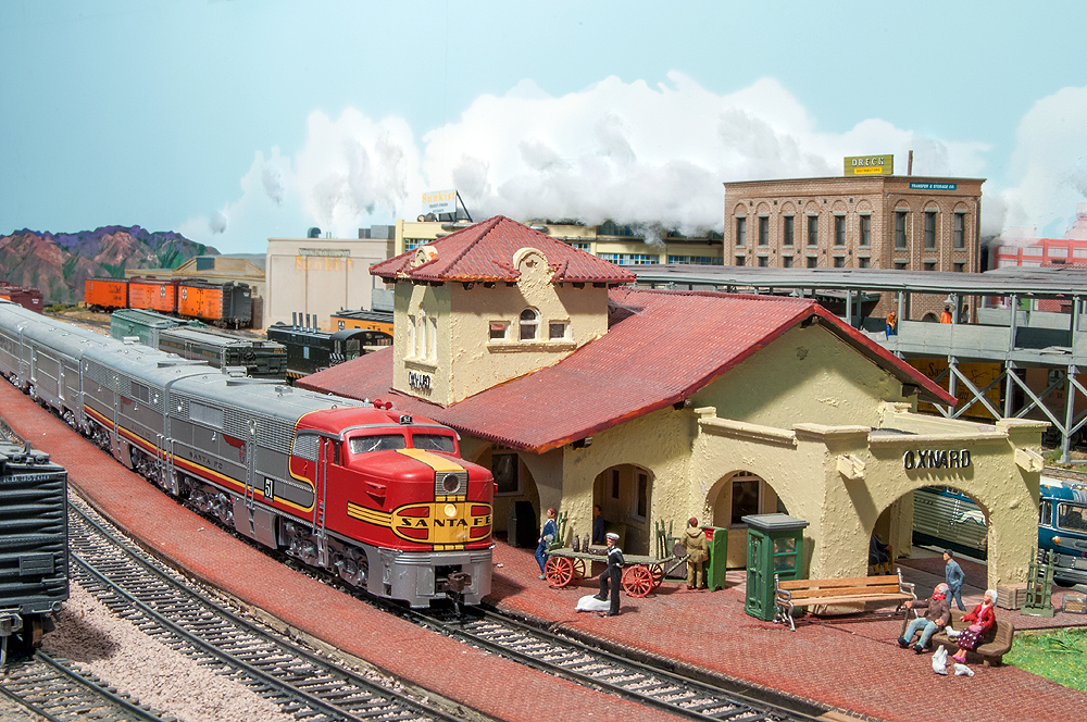 A red-and-silver Santa Fe diesel locomotive leads a train paused at a southern California city.