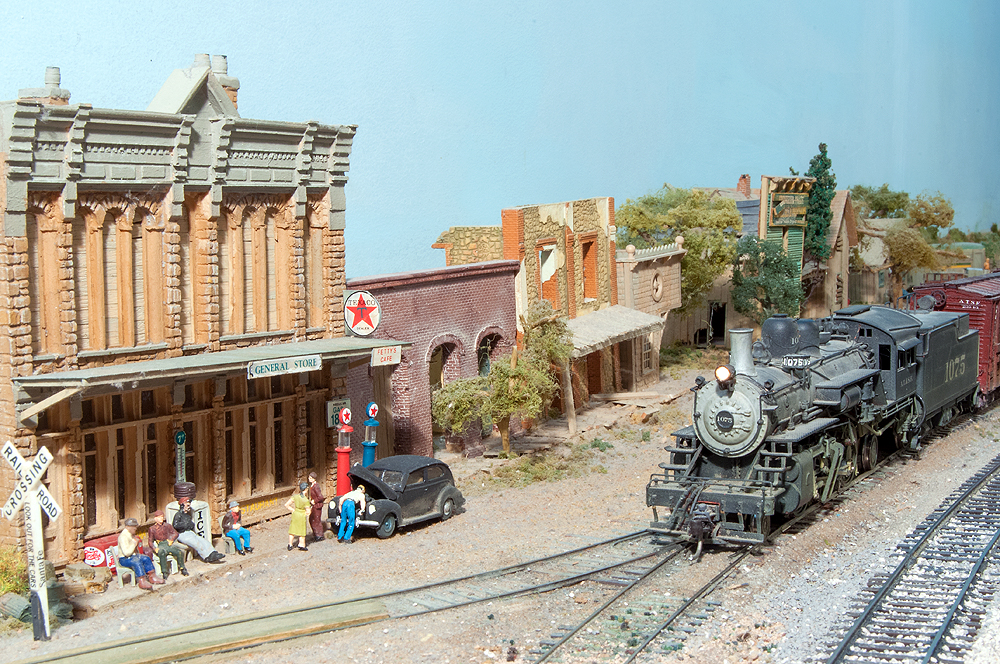 2-6-2 steam locomotives with freight train trundles down dirt-covered tracks in a small-town Western scene.
