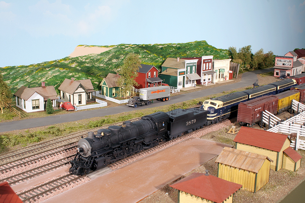 HO Scale Santa Fe steam and diesel locomotives team up to pull a train through a small town scene.