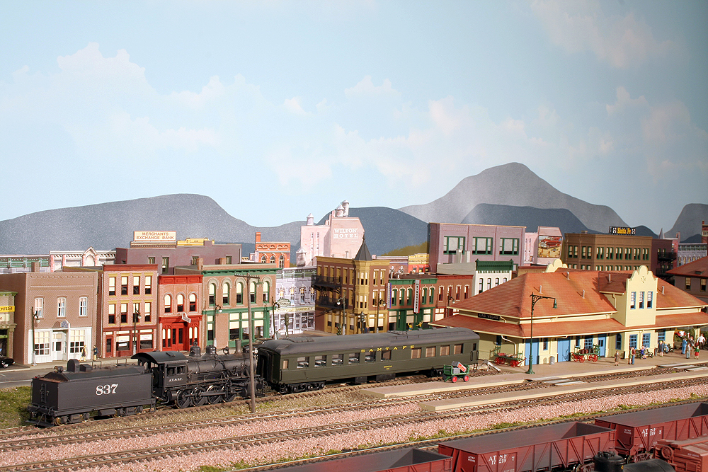 A small consolidation-type locomotive pushes a diner to a bumper post next to a Santa Fe depot in a yard with at least six parallel tracks.