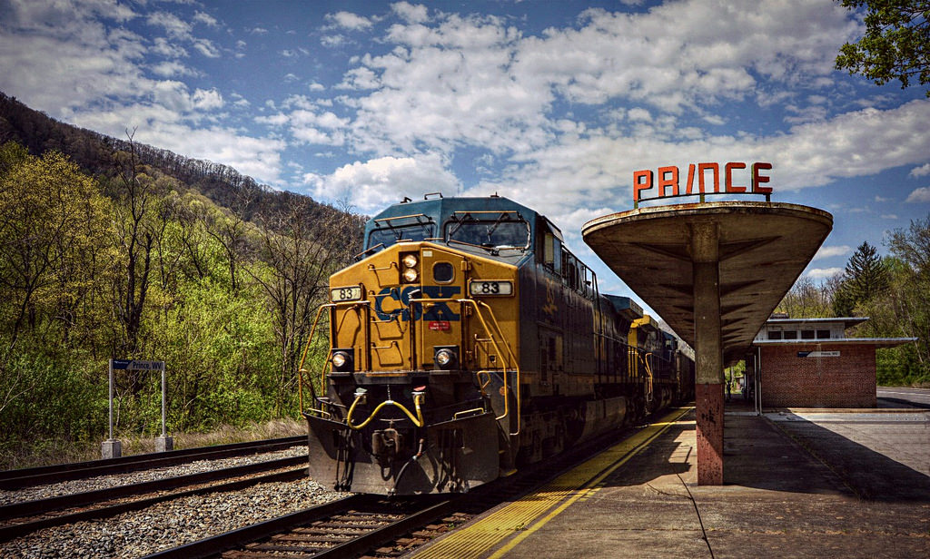 A yellow-and-blue locomotive leads a freight train beside a passenger platform bearing the word "Prince".