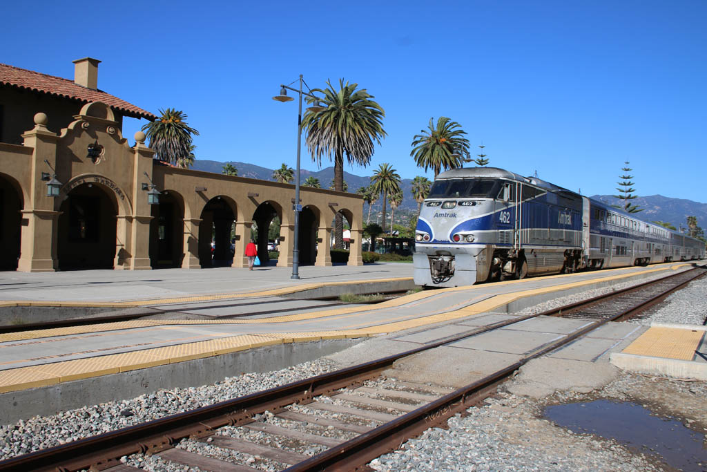 An Amtrak train paused at an adobe-style train station under clear skies with palm trees.
