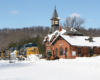 A blue-and-yellow diesel locomotive leads a freight train past a gothic revival train station in snow.