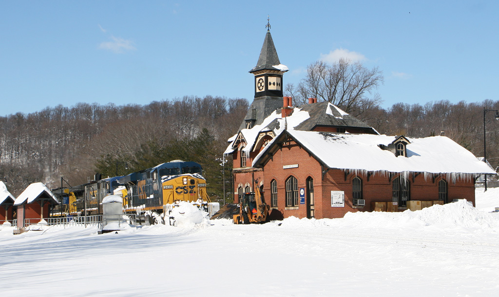 A blue-and-yellow diesel locomotive leads a freight train past a gothic revival train station in snow.