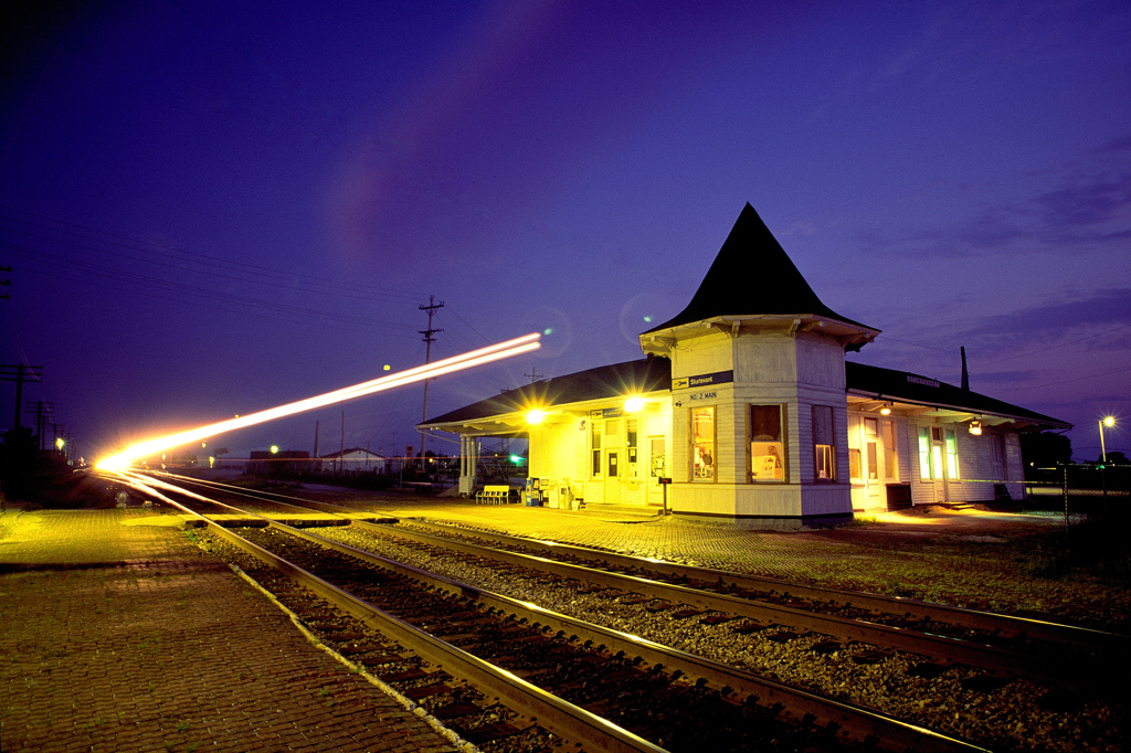 A light blur appears in front of an illuminated passenger train station in the evening.
