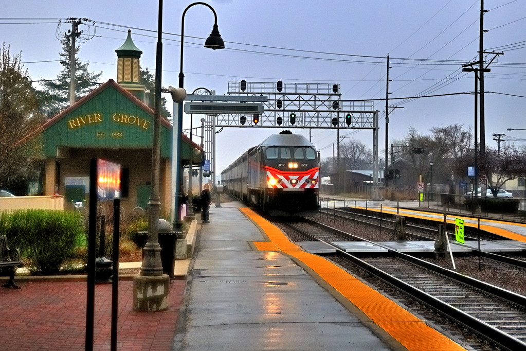 A stripped-nose locomotive pull a passenger train in rain by a small brick train station.