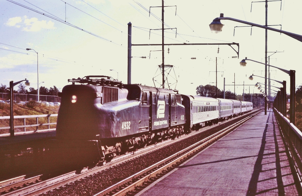 A streamlined electric locomotive hauling a passenger train beside a passenger platform.
