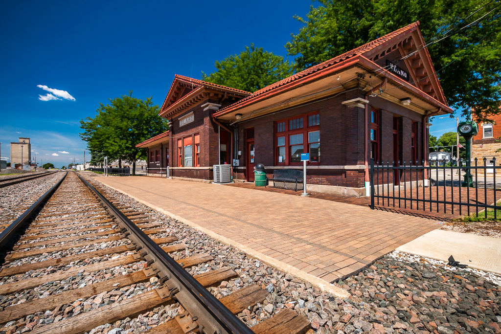 A one-story brick passenger train station appears in sunlight under clear skies in summertime.