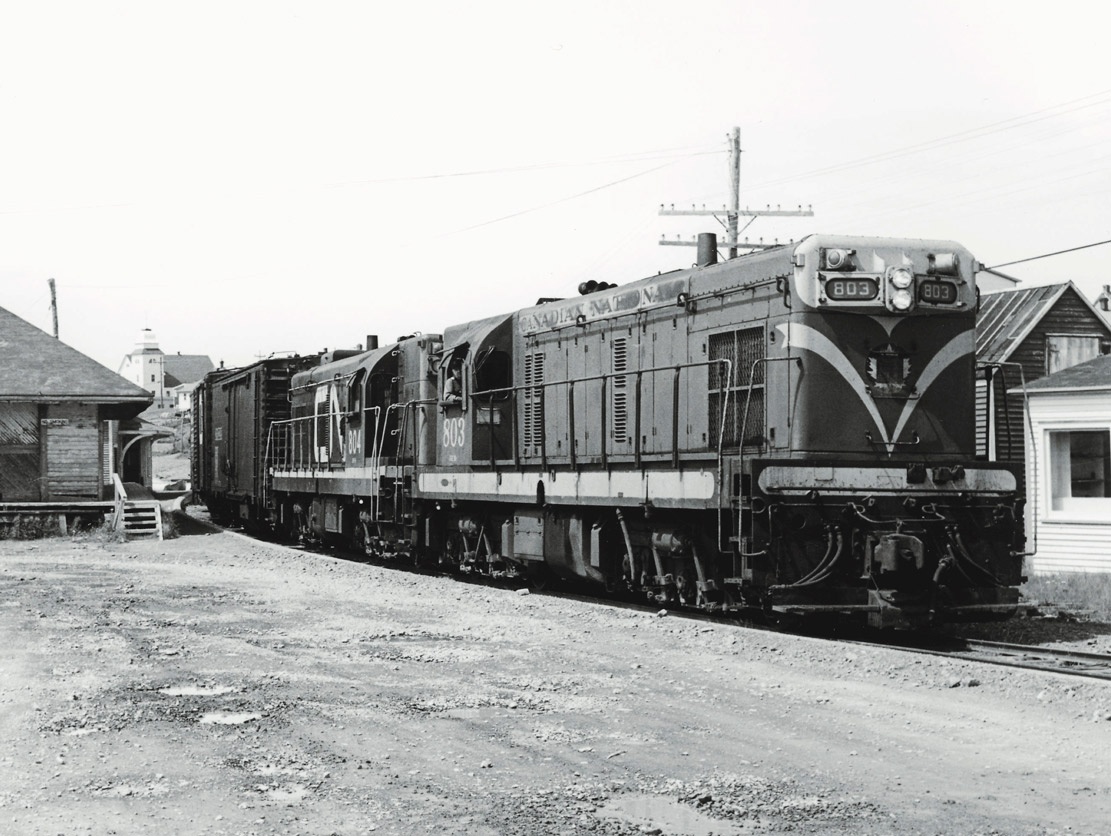 Two diesel electric locomotives at the head of a train amid a handful of age-worn buildings.