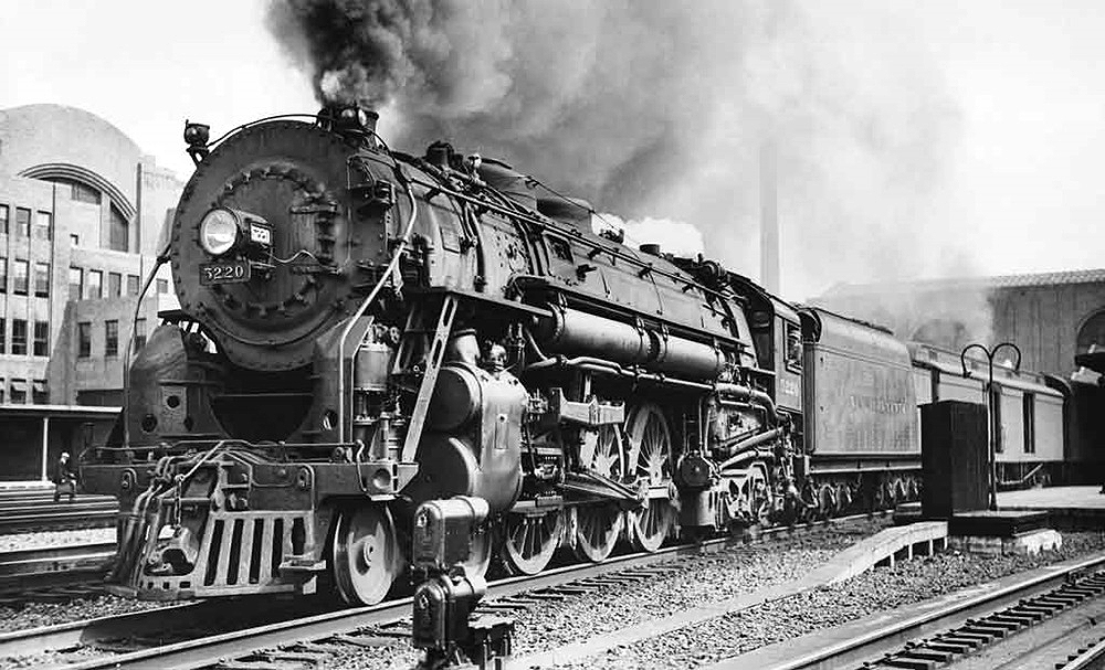 A steam locomotive appears with a train in a rail yard.