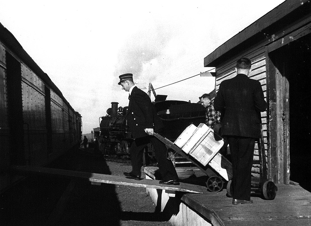 Uniformed railroad worker pulls a loaded dolly up a ramp to an express car.