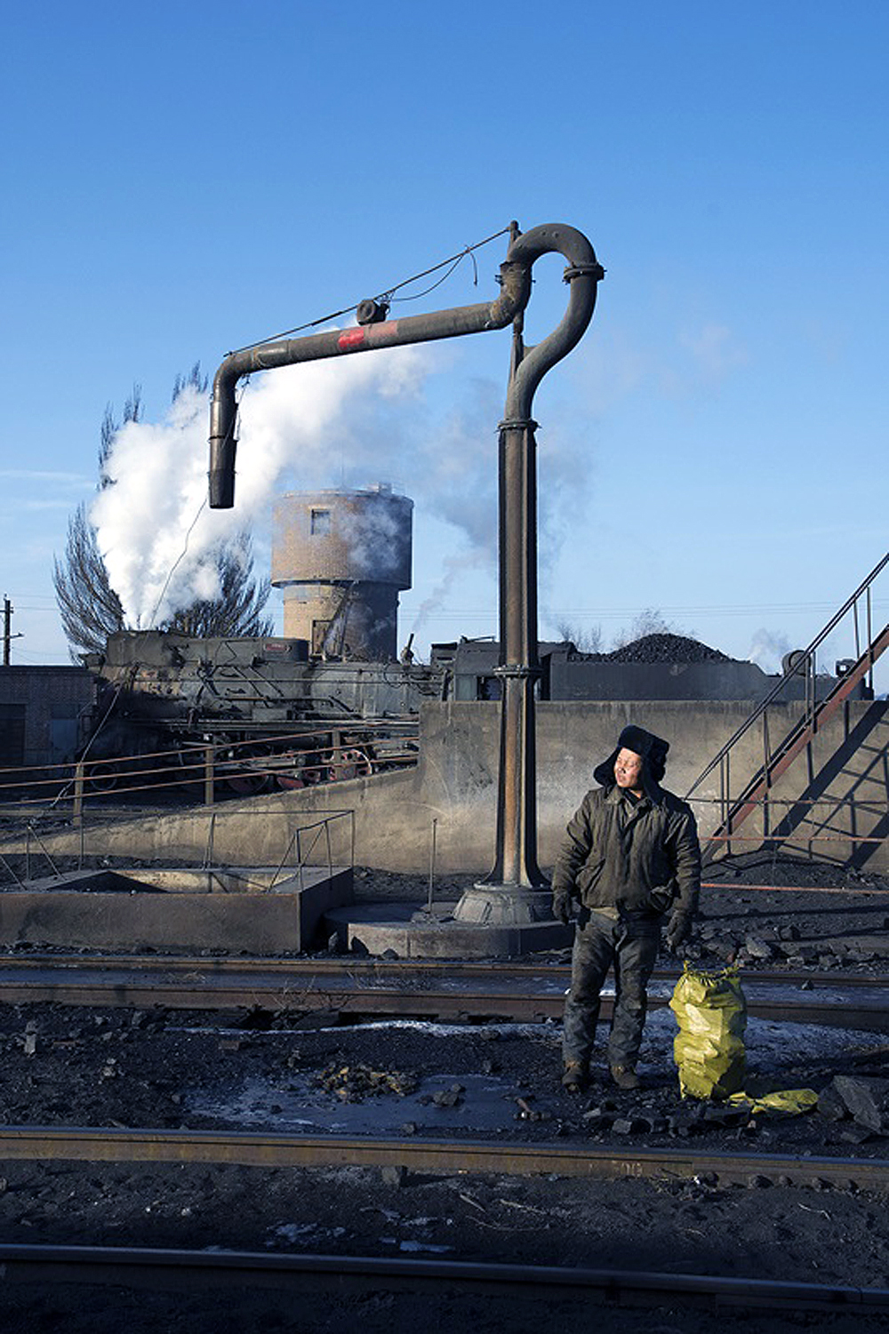 A man stands near a water standpipe in a rail yard.