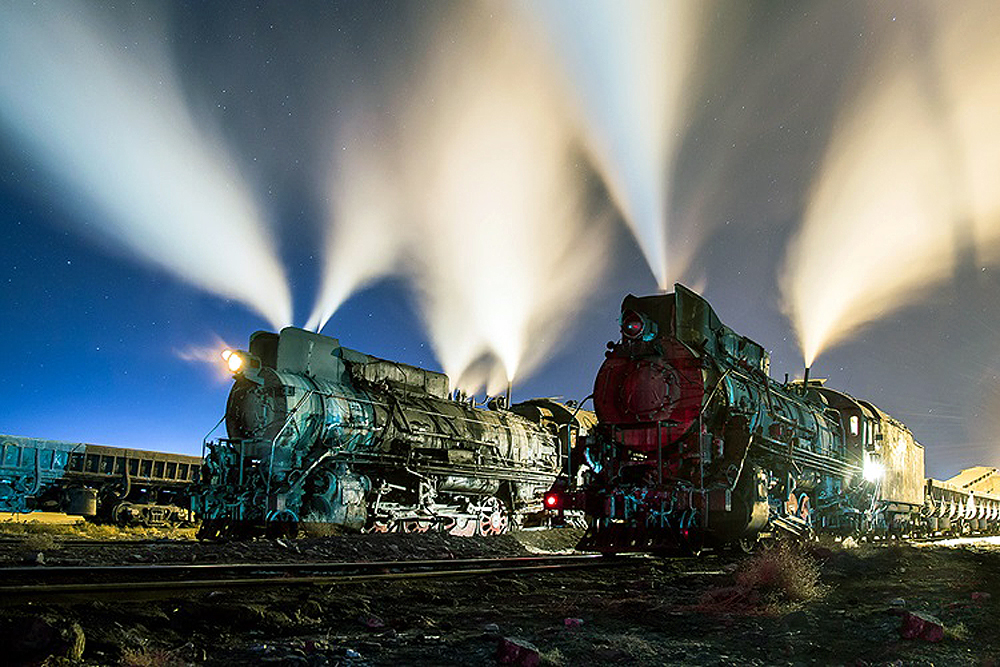 Two steam locomotives emitting steam on a star-lit night in a rail yard.