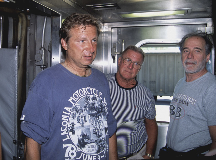 Three men in summer wear standing and listening.