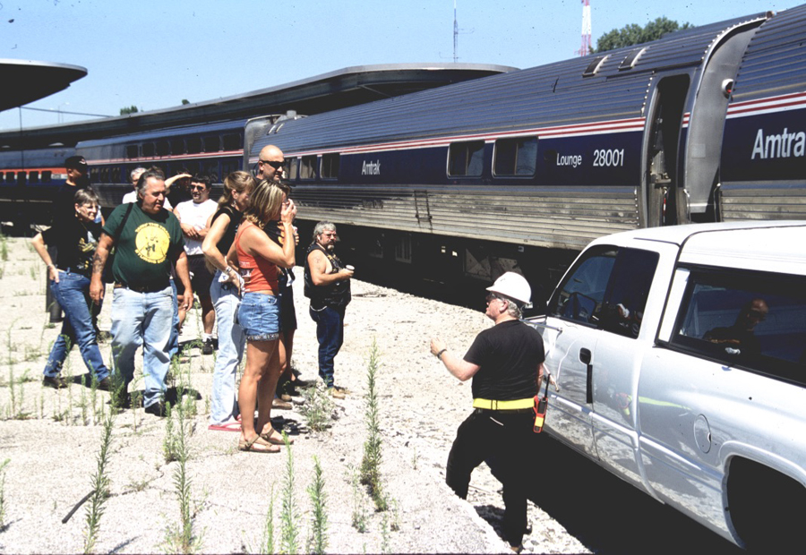 A helmeted worker speaks to gathered people in front of a train.