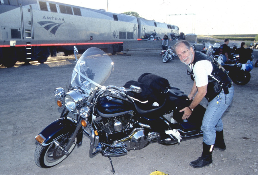 Man posing with motorcycle near a passenger train.