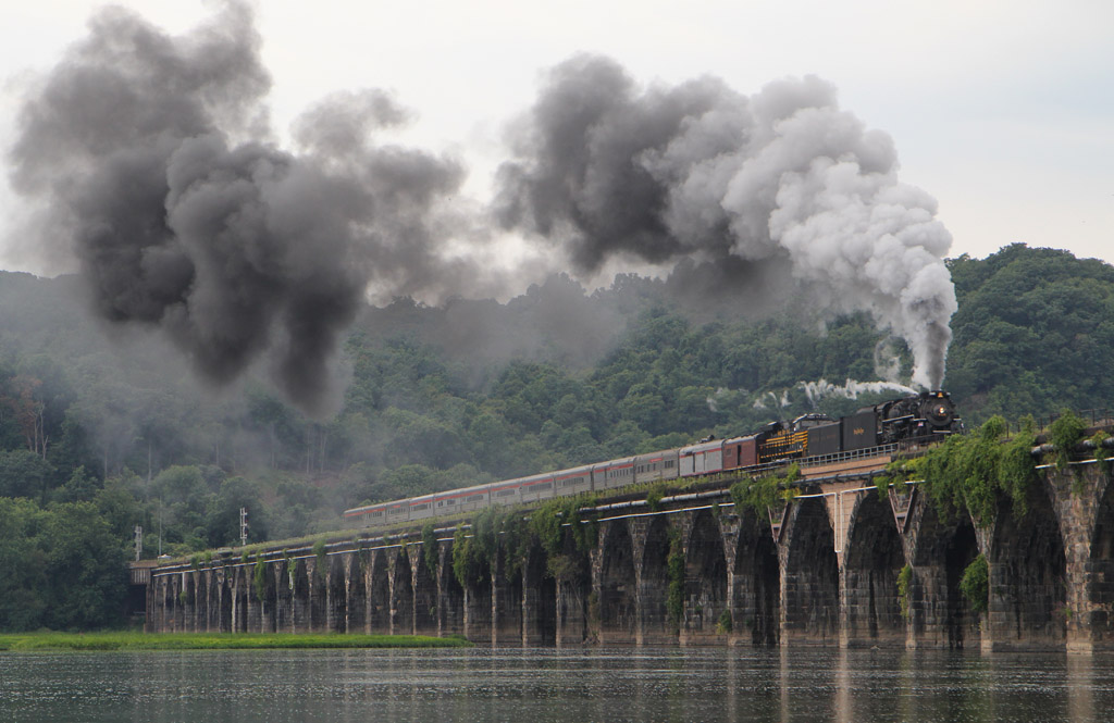 Steam locomotive leading a passenger train over a river on an old stone bridge.