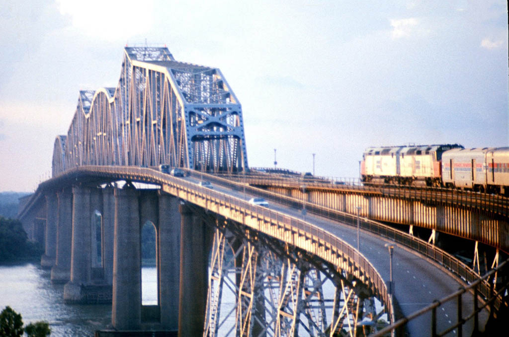 Amtrak diesel locomotives lead a passenger train over a large truss span bridge over a river.